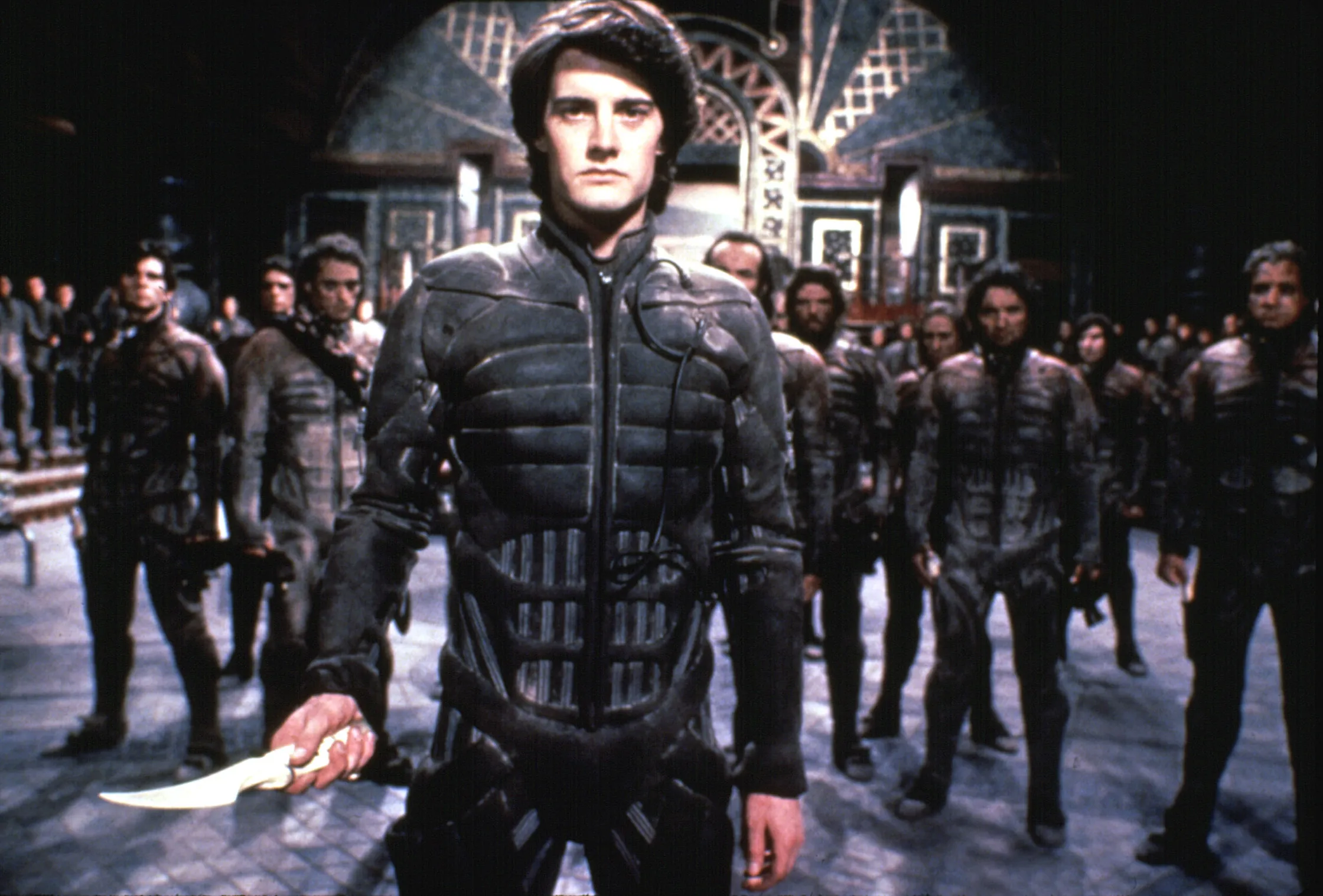 Person wearing black padded suit holding a knife while a crowd of people stand behind them