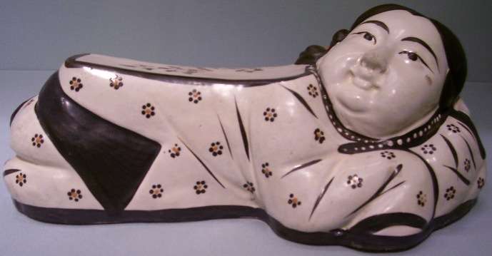“Pillow in the form of a reclining girl, Northern China, Jin dynasty (1115-1234), Cizhou ware, high-fired ceramic with overglazed decoration, The Avery Brundage Collection, B60P422. The representation of girls on a Song pillow, rare in contrast to the frequent representations of boys, suggests a piece made for female use. This finely sculpted pillow features a young girl, her expression well detailed, accompanied by chrysanthemum and plum blossoms.”